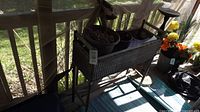 Wicker planter box elevated on wooden legs with several plant pots placed inside, viewed from the front with some outdoor light and background plants.