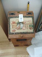Front view of wooden potato and onion bin with hinged top featuring painted baskets of potatoes and carrots, and a knob for opening.