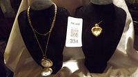 Two necklaces displayed on black busts: one gold plated pocket watch on chain with flip-open cover, the other a gold plated heart-shaped watch pendant.