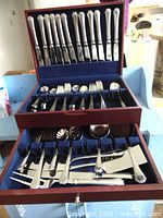 Front view showing two-tier wooden box containing multiple stainless steel cutlery pieces; knives in upper tier, forks and spoons in lower tier with blue cloth lining.