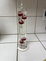 Full view of the 14.5-inch Galileo thermometer standing upright showing red colored glass balls floating in clear liquid inside a cylindrical glass tube.