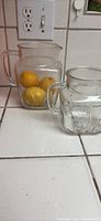 Photo showing two clear glass pitchers with starburst patterns on a white tiled counter, one taller containing lemons, the other shorter.
