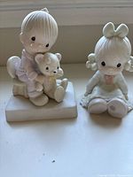 Front view of two porcelain Precious Moments figurines. Left is a girl holding a teddy bear seated on a rectangular base. Right is a girl sitting on the floor with a large bow on her head.