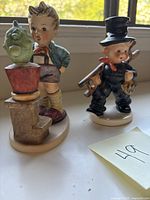 Two Goebel figurines side by side on a window sill, one of a boy with a cactus plant and one chimney sweep with ladder.