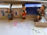 Three Hummel figurines displayed with original boxes and display cards on a shelf.
