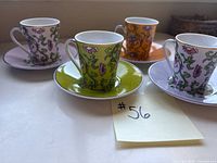 Four espresso cups with matching saucers displayed together; cups in orange, green, and two light lavender bases featuring floral patterns with pink and white flowers and green vines, white interiors, and purple handles.