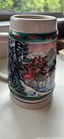 Side view of the ceramic beer stein showing winter holiday scene with horse-drawn carriage, snow-covered pine trees, and pine cones.
