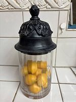 Front view of the glass jar filled with lemons, showing black ornate lid with detailed leaf design and finial handle.