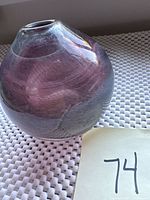 Clear glass vase with purple and white swirls, round shape with narrow opening, artist-signed, displayed on a white textured surface with a handwritten '74' tag.