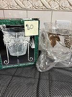 Clear crackled glass ice or plant bucket with silver plated stand featuring grapevine decorations next to its box.