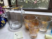 Photo showing the complete set: one clear glass ice/champagne bucket with gold band and three small glasses with gold patterning arranged on a window ledge