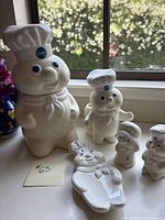 Photo of entire Pillsbury Doughboy collectible set showing cookie jar, salt & pepper shakers, spoon rest, and vase on window ledge with natural light and floral background.