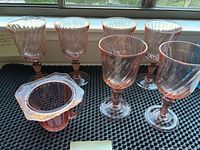 Set of six pink Depression glass wine glasses with a French swirl pattern around the bowl, arranged on a black mat near a window. Included is a sugar bowl also made of pink Depression glass.
