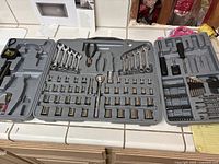 Full view of organized plastic tool case with wrench set, socket set, ratchets, wire cutters, drill bits, Allen wrenches, and screwdrivers. Several empty slots show missing tools. Case lid closes but with loose door attachments.