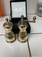 Two silver-tone salt and pepper shakers, one with grinder handle, placed in front of open green box.