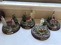 Four Thomas Kinkade collectible cottages with lighthouses arranged on a window ledge showing details and scale.