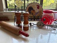 Photo showing a wooden rolling pin with a red handle tip, a pair of wooden salt and pepper grinders with clear centers, a decorative ceramic plate with a rooster design on a metal wire rack, a red glass popcorn popper, and part of a wood handle crank mixer.