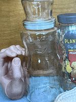 Close-up image of the clear glass jar in the shape of Mr. Peanut, held by hand showing facial details and separate hat-shaped lid.