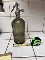 Full image of the greenish tinted glass bottle with metal top, shows measuring tape at base indicating about 4 inches in width.