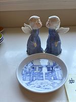 Two porcelain figurines of kissing angels in blue dresses behind a small blue and white porcelain dish on a window sill.