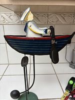 Front and right side view of the vintage metal sailor in a rowing boat balance toy showing the painted details and the black paddles on metal stand.