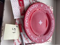 Photo showing red ceramic plate with white lettering on the rim, a red pen, packaging paper with red logos, and a handwritten number tag.