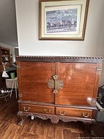Front view showing double doors with large decorative metal lock plates, base with detailed carving and two drawers with brass handles.