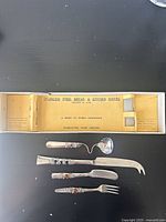 Silverware cutlery set including bread and kitchen knives, fork, and ladle laid out next to original box open showing yellow vintage print label.