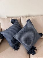 Two blue striped square throw cushions with tassels on corners placed on a light beige couch.