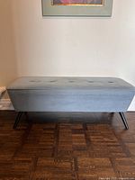 Front view of blue velvet bench showing entire top and legs, set against a wall with hardwood flooring below.