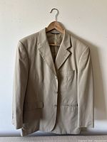 Front view of beige Brooks Brothers suit jacket hanging on a wooden hanger against a white wall showing the three-button front, notch lapels, chest pocket, and flap pockets.