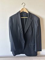 Front view of dark charcoal Brooks Brothers suit jacket hanging on wooden hanger against white wall showing notch lapels and two front buttons