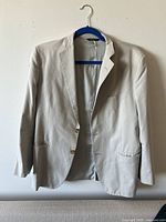 Front view of light beige single-breasted suit jacket with two-button closure and flap pockets, hanging on blue hanger