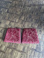 Top view of two trapezoid chair seat covers with red fabric featuring cream swirls, placed on wooden floor.