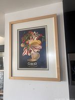 Full view of framed art print hanging on white wall, showing the woman and Can CIRIO image with wooden frame.