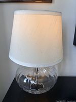 Glass desk lamp showing round clear glass base and cream/off-white fabric lampshade, lit to show soft ambient light.