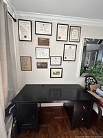 Front view of a large black wooden desk under a wall with framed certificates, showing top surface and storage compartments.