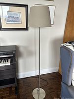 Full view of tall standing floor lamp near piano and chair showing beige lampshade and metal pole base.