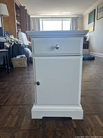 Front view of the white bedside table showing a single drawer on top with round silver knob and a cupboard door below with matching silver knob hardware, standing on dark wooden floor.