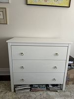Front view of the white chest of drawers showing three drawers with gold ring pull hardware on a white flat background.