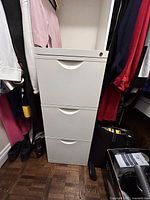 White three drawer filing cabinet positioned between hanging clothes. Top drawer has a lock and curved recessed handles on all drawers.