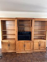 Three-piece wood furniture with two bookcases flanking a central display cabinet, finished in warm tones with visible hardware on lower cabinets. Central cabinet shows a TV on shelf with two shelves above and lower closed compartments with missing hardware handles.