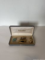 Photo showing a Richard Booth box open with the set inside, including a small blue pin, a pearl Miriam Haskell brooch, a gold-tone decorative brooch, and the pocket watch.