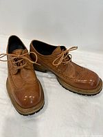 Pair of caramel brown leather brogue shoes with lace-up closure placed side by side on white surface. Shows detailed perforation and leather texture.