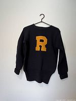 Full front view of navy sweater with yellow 'R' patch hung on hanger against plain wall