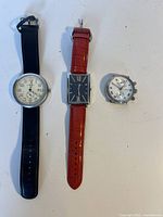 Four wristwatches arranged on a white surface: one round with white dial and black strap, one rectangular with black dial and red strap, two round silver-tone watches with no straps.
