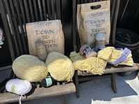 Full view of yarn skeins in various colors with two Kertzer 'Down to Earth Cotton' packaging bags and partial knitting samples on wooden chairs outdoors.