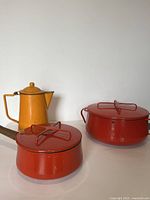 Front view showing small red saucepan with wooden handle, large red Dutch oven, and orange coffee pot