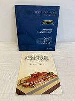 Blue hardcover pop-up book titled 'Frank Lloyd Wright In Pop-Up' and a booklet for the Robie House model kit with a colored illustration on the cover.