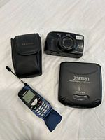 Photo showing Sony Discman, Samsung flip phone, and Pentax camera with case on white fabric background.